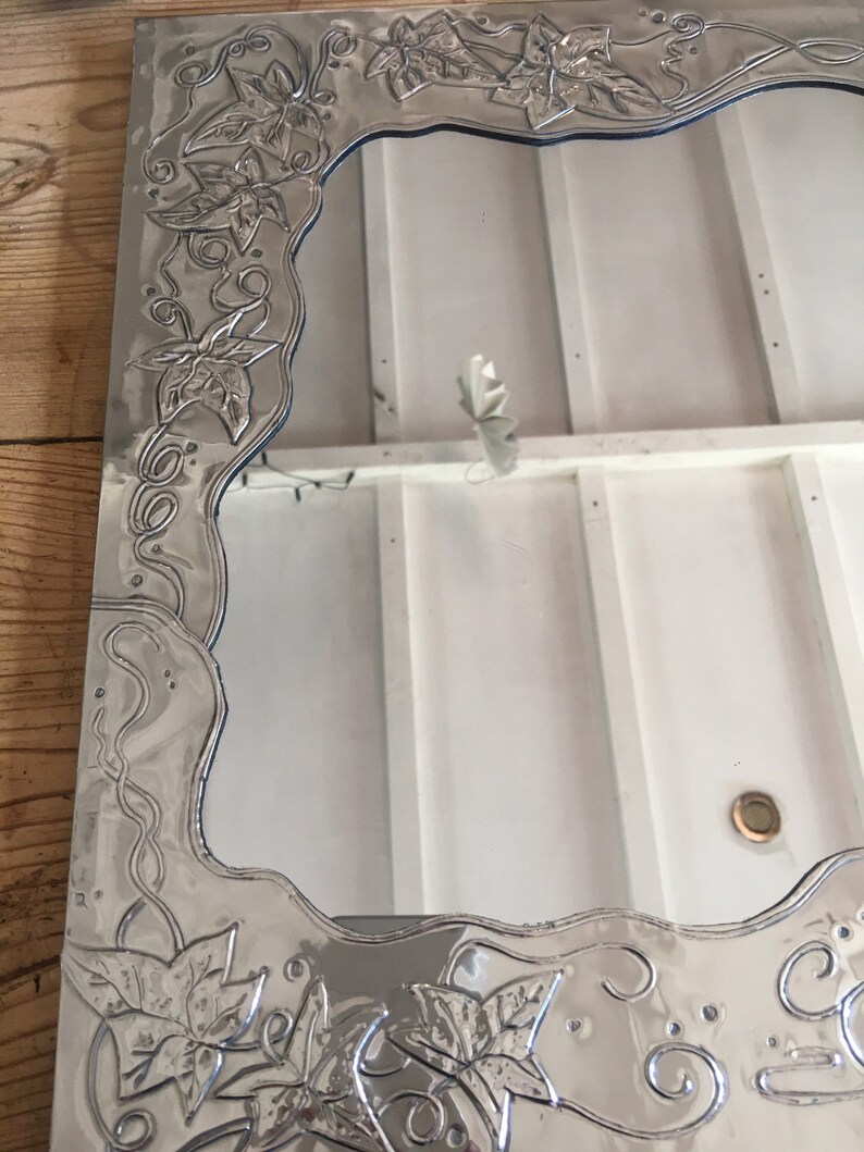 May include: Silver-toned rectangular mirror with an ornate, embossed border featuring a repeating leaf and vine design. The mirror's frame has a wavy edge and a reflective surface, suitable for home decor.