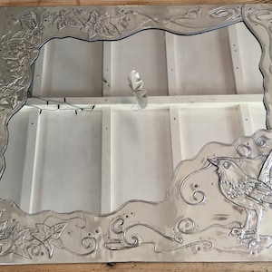 May include: A silver-colored metal mirror frame with an embossed design. The frame features a bird, leaves, and swirling patterns. The mirror's interior is white, and the frame is rectangular.