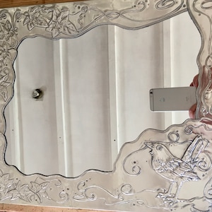May include: Silver-toned rectangular mirror with an ornate, embossed frame. The frame features a bird and leaf design. The mirror reflects a white wall and a phone. The mirror is a decorative home decor item.