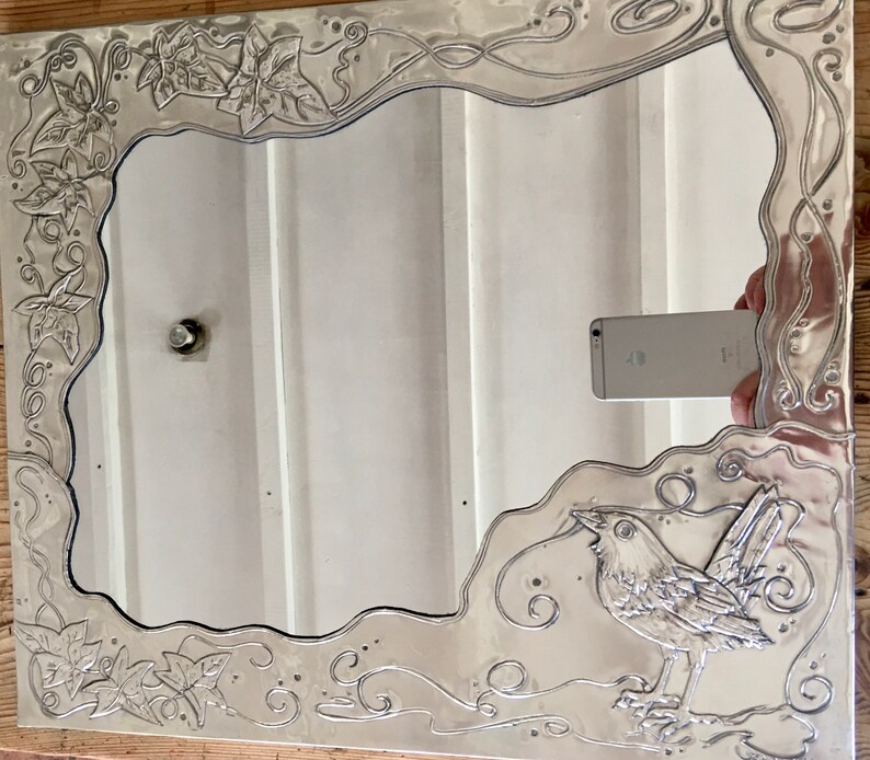 May include: Silver-toned rectangular mirror with an ornate, embossed frame. The frame features a bird and leaf design. The mirror reflects a white wall and a phone. The mirror is a decorative home decor item.
