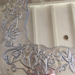 May include: A silver-colored, decorative picture frame with embossed leaf and vine designs. The frame has a wavy edge and features detailed botanical patterns, adding a touch of nature-inspired elegance to home decor.