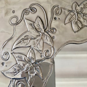 May include: Close-up of a silver-colored metal decorative piece with embossed leaf and vine designs. The leaves have detailed veining, and the vines feature delicate curls. The piece has a reflective, polished finish, and the design is raised from the surface.