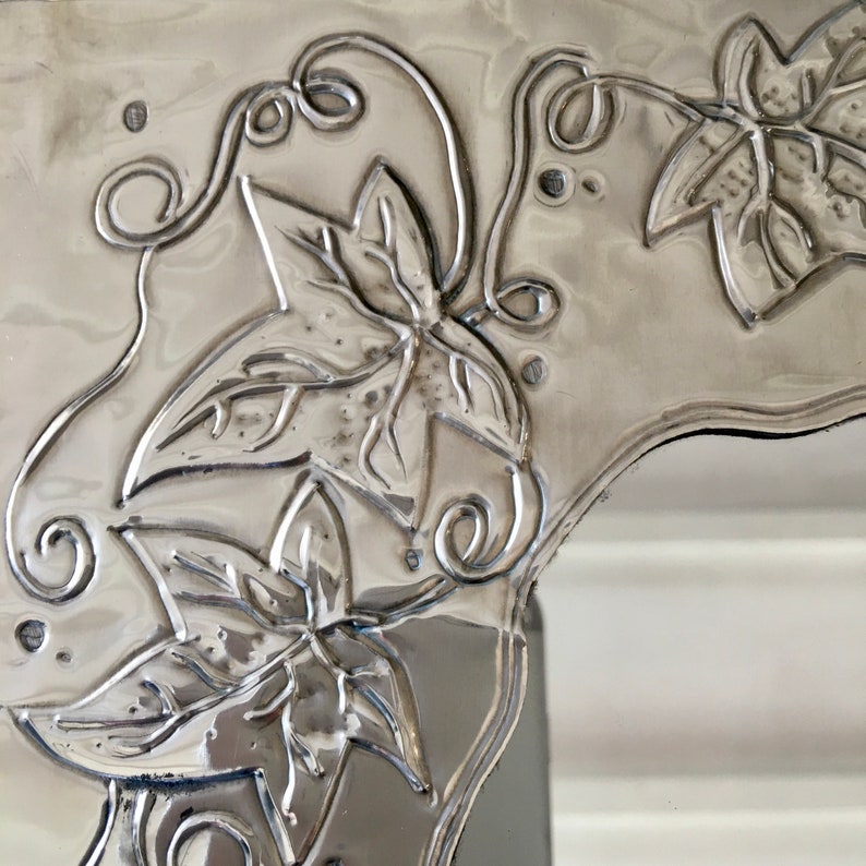 May include: Close-up of a silver-colored metal decorative piece with embossed leaf and vine designs. The leaves have detailed veining, and the vines feature delicate curls. The piece has a reflective, polished finish, and the design is raised from the surface.