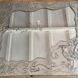 May include: A silver-colored metal mirror frame with an embossed design. The frame features a bird, leaves, and swirling patterns. The mirror's interior is white, and the frame is rectangular.