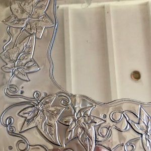May include: A silver-colored mirror with a decorative border featuring embossed leaf and vine designs. The leaves have detailed veining, and the border has a wavy edge. The mirror is set against a white background.