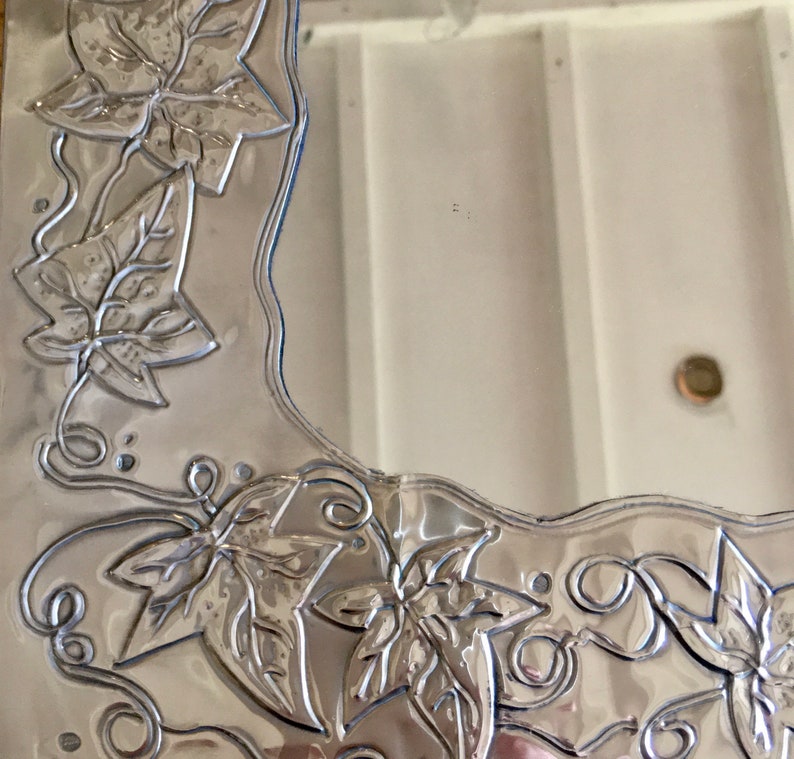 May include: A silver-colored mirror with a decorative border featuring embossed leaf and vine designs. The leaves have detailed veining, and the border has a wavy edge. The mirror is set against a white background.