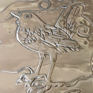 May include: A detailed, silver-colored relief of a bird perched on a branch. The bird is depicted with intricate feather details and stands out against the textured background. The artwork showcases a nature-inspired theme.