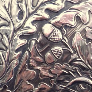 May include: Close-up of a silver embossed design featuring oak leaves and acorns. The design is detailed and intricate, with a high-polish finish.