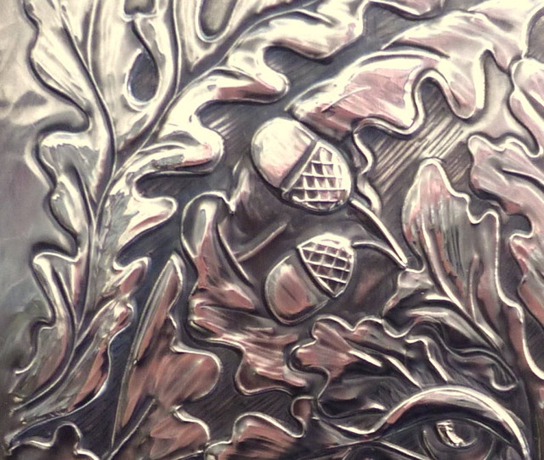 May include: Close-up of a silver embossed design featuring oak leaves and acorns. The design is detailed and intricate, with a high-polish finish.