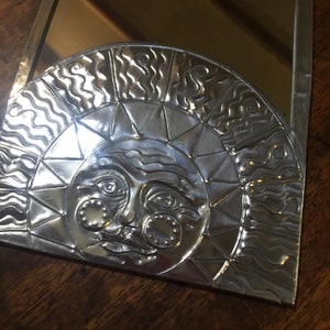 May include: A silver metal mirror with a sun face design. The sun has a face with eyes, a nose, and a mouth. The sun is surrounded by a circle of wavy lines.