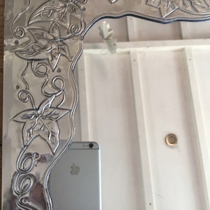 May include: A silver-colored mirror with an embossed floral design. The mirror's frame features detailed leaves and vines, creating a decorative border. The design is raised, adding texture to the metallic surface. The mirror reflects a room.