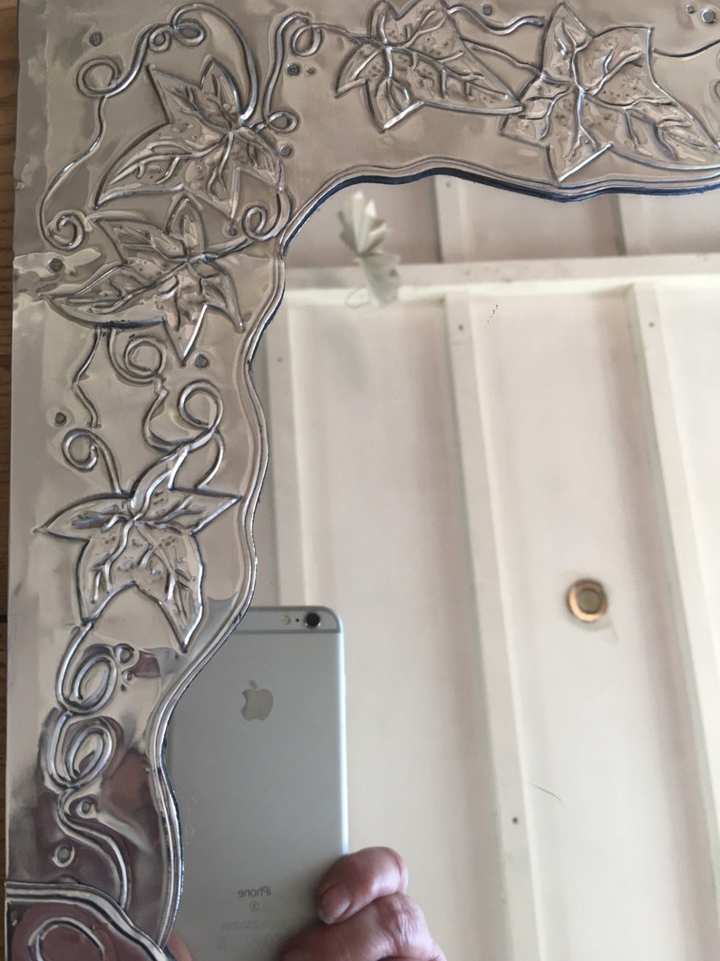 May include: A silver-colored mirror with an embossed floral design. The mirror's frame features detailed leaves and vines, creating a decorative border. The design is raised, adding texture to the metallic surface. The mirror reflects a room.