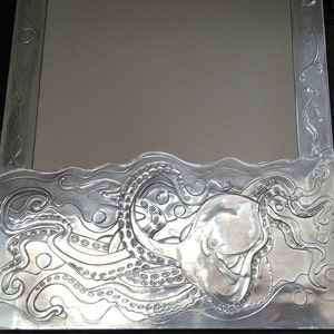 May include: A silver mirror with an embossed octopus design around the frame. The octopus is surrounded by waves and bubbles.