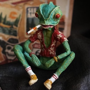 May include: A handmade green chameleon figurine wearing a red floral shirt, white socks with red stripes, and yellow shoes, holding a cigarette. The figurine is seated with a backdrop of vintage-style travel stickers and an American flag.