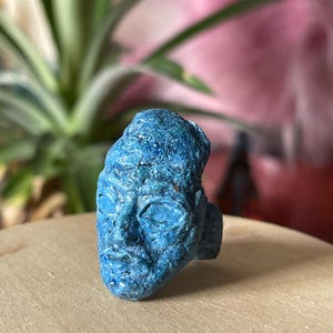 Puede incluir: Un anillo que presenta un rostro azul esculpido con un acabado mate y texturizado. El diseño muestra una cabeza de aspecto humano con ojos cerrados, nariz y labios definidos, elaborado en un tono azul monocromático con irregularidades visibles.