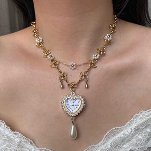 Puede incluir: Collar dorado con un colgante de reloj en forma de coraz&oacute;n. El reloj tiene una esfera blanca con n&uacute;meros negros y un borde de cristal. El collar incluye detalles florales y perlas, y una gota de perla en forma de pera.