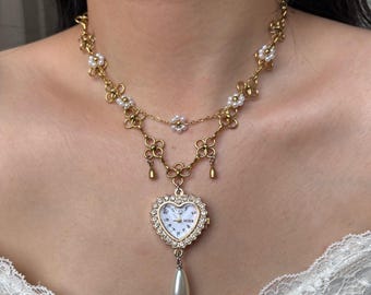 May include: Gold-toned necklace featuring a heart-shaped watch pendant. The watch has a white face with black numerals and is surrounded by a crystal-like border. The necklace includes floral and pearl accents, and a pear-shaped pearl drop.