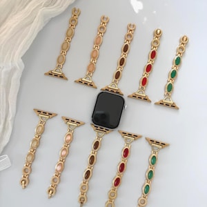 May include: Gold-toned watch bands with oval-shaped colored accents. The bands are in various colors, including beige, brown, red, and green. The bands are designed to fit a smartwatch.