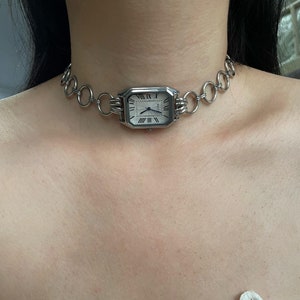 Puede incluir: Un collar choker plateado con una esfera de reloj cuadrada con números romanos. La esfera del reloj está unida a la cadena con dos eslabones plateados.