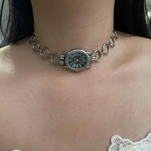 May include: A silver chain choker necklace with a round watch face attached. The watch face has black Roman numeral markings and a black background.
