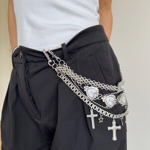 May include: Silver chain belt with multiple strands, heart-shaped charms, crosses, and a star. The belt is attached to black pants. The model is wearing a white tank top.