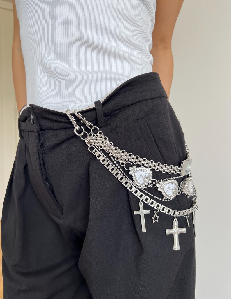 May include: Silver chain belt with multiple strands, heart-shaped charms, crosses, and a star. The belt is attached to black pants. The model is wearing a white tank top.