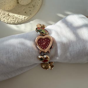 May include: A heart-shaped wristwatch with a red face and gold-tone accents. The watch features decorative charms with floral and berry designs. The band is adorned with additional charms. The watch is approximately 18 cm in length.