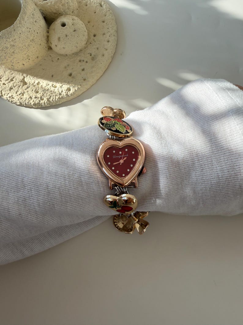 May include: A heart-shaped wristwatch with a red face and gold-tone accents. The watch features decorative charms with floral and berry designs. The band is adorned with additional charms. The watch is approximately 18 cm in length.