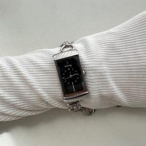 May include: A rectangular silver-tone wristwatch with a black dial featuring the text GoGoey. The watch has a metallic link bracelet with decorative flower-shaped accents and a crown on the right side, displayed on a white ribbed fabric sleeve.