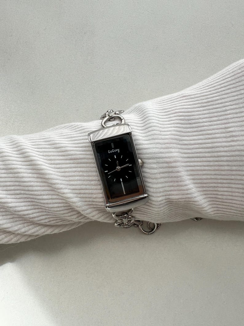 May include: A rectangular silver-tone wristwatch with a black dial featuring the text GoGoey. The watch has a metallic link bracelet with decorative flower-shaped accents and a crown on the right side, displayed on a white ribbed fabric sleeve.