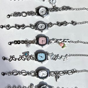 May include: A collection of seven silver-toned wristwatches with various face shapes, including round, square, and oval. Each watch features a different style of band, some with decorative charms like flowers, hearts, and snowflakes. The watch faces display traditional analog time.