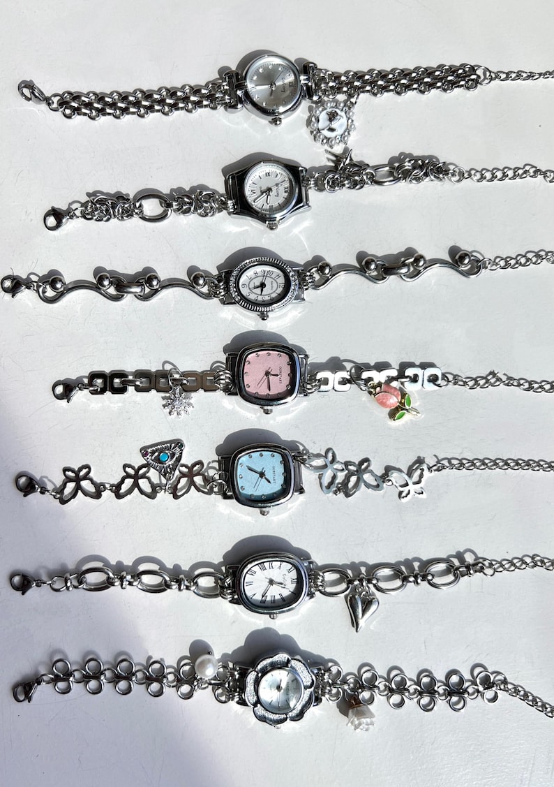 May include: A collection of seven silver-toned wristwatches with various face shapes, including round, square, and oval. Each watch features a different style of band, some with decorative charms like flowers, hearts, and snowflakes. The watch faces display traditional analog time.