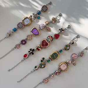 May include: Five decorative wristwatches with silver chains and colourful, ornate faces. Each watch features a unique design, including floral, heart, and geometric shapes, with various coloured gemstones. The watch faces are in shades of gold, purple, green, and pink.