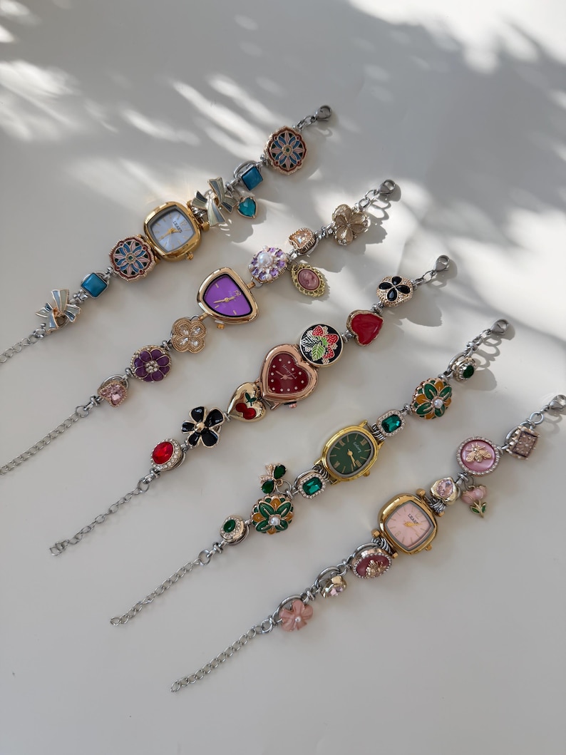 May include: Five decorative wristwatches with silver chains and colourful, ornate faces. Each watch features a unique design, including floral, heart, and geometric shapes, with various coloured gemstones. The watch faces are in shades of gold, purple, green, and pink.