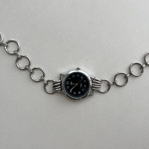 May include: A silver chain watch with a black face and silver hands. The watch is attached to a silver chain with circular links.