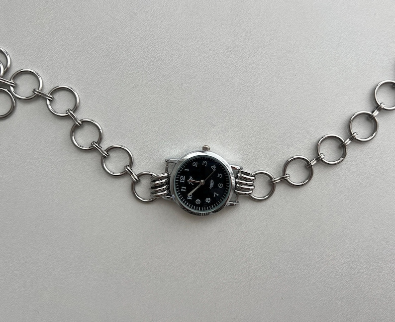 May include: A silver chain watch with a black face and silver hands. The watch is attached to a silver chain with circular links.