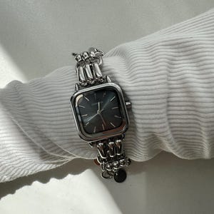 May include: A silver-toned wristwatch featuring a square case with a dark gray dial and the brand name JENISSAN printed in white. The watch has a metallic link bracelet composed of rounded, interconnected segments and a small circular charm.