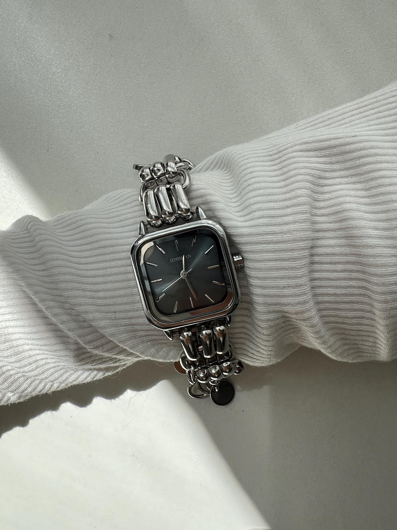 May include: A silver-toned wristwatch featuring a square case with a dark gray dial and the brand name JENISSAN printed in white. The watch has a metallic link bracelet composed of rounded, interconnected segments and a small circular charm.