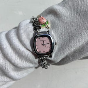 May include: A silver-toned wristwatch with a square pink face and a silver chain band. The watch face has small crystals and the text "OURSTART". A charm of a pink rose with green leaves hangs from the band.
