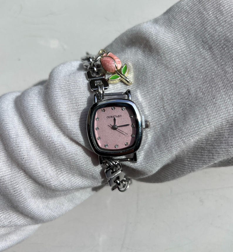 May include: A silver-toned wristwatch with a square pink face and a silver chain band. The watch face has small crystals and the text "OURSTART". A charm of a pink rose with green leaves hangs from the band.