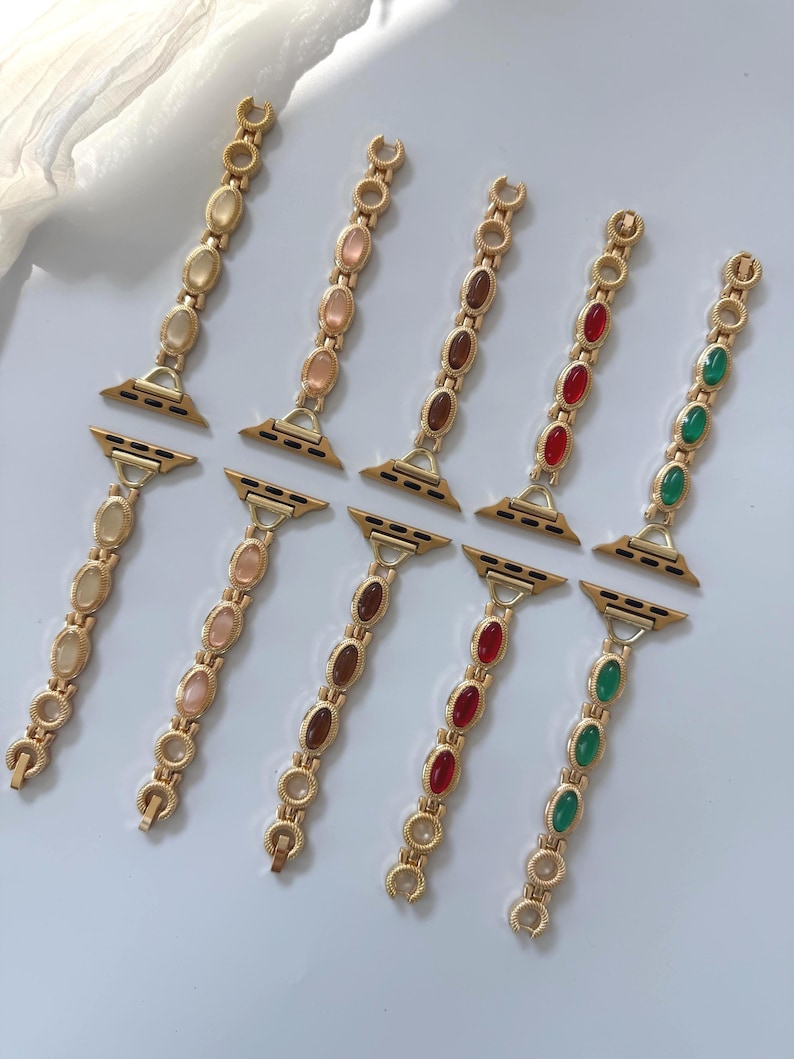 May include: Gold-toned watch bands with oval-shaped gemstones in various colors, including yellow, pink, brown, red, and green. The bands are designed to attach to a smartwatch and feature a decorative chain-link style.
