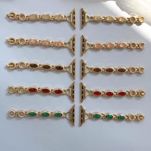 May include: Gold-toned watch bands with oval-shaped gemstones in various colors: cream, pink, brown, red, and green. The bands are designed to attach to a smartwatch. The bands are displayed on a white surface.