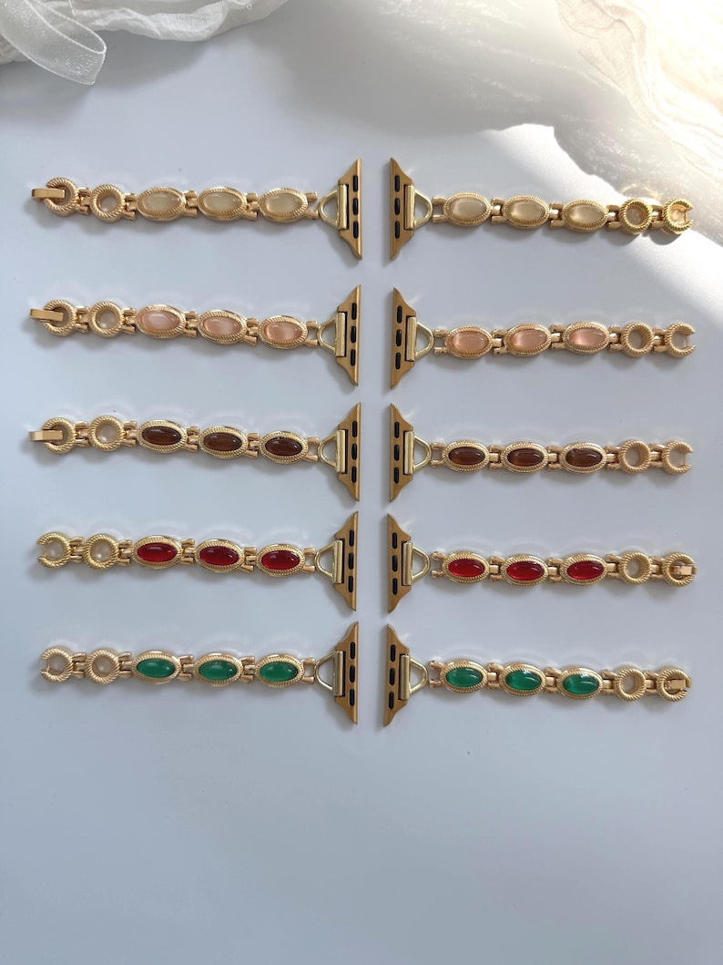May include: Gold-toned watch bands with oval-shaped gemstones in various colors: cream, pink, brown, red, and green. The bands are designed to attach to a smartwatch. The bands are displayed on a white surface.