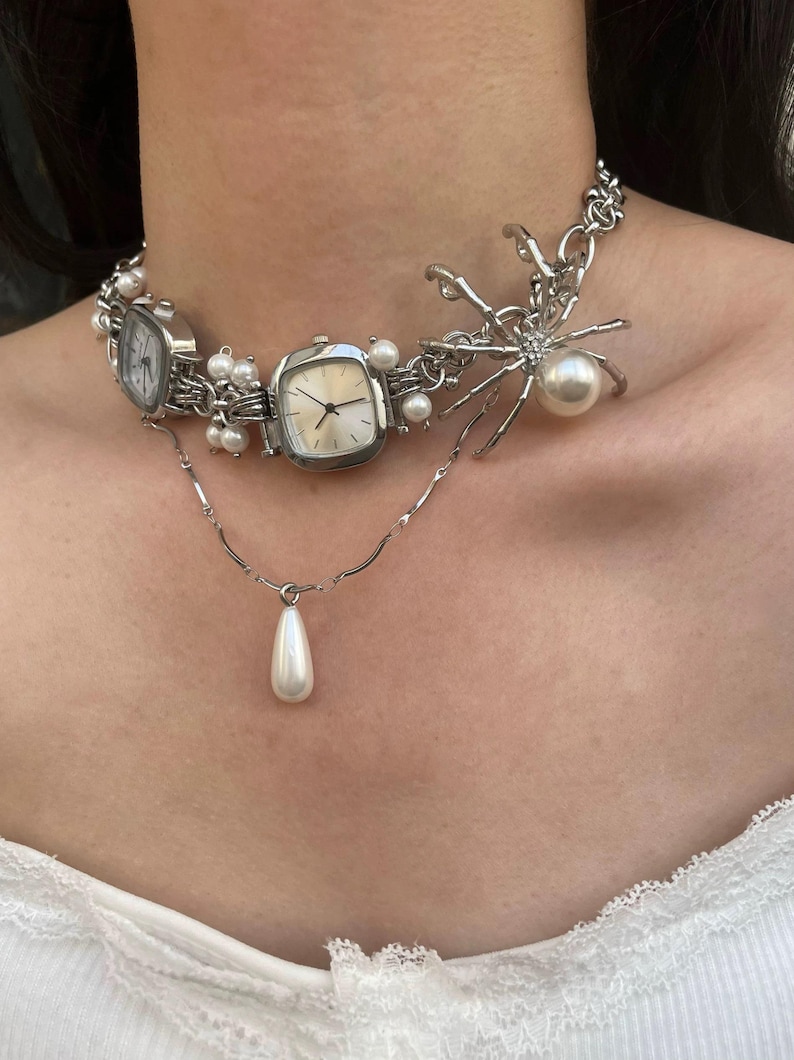 May include: A silver choker necklace with multiple watch faces, pearls, and a spider charm. The spider charm has a large pearl in the center.