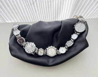 May include: Black leather handbag with a silver watch strap handle. The handle is composed of various silver watches with white faces and different shaped dials. The bag is gathered at the top and has a soft, slouchy appearance.