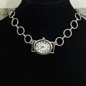 May include: A silver chain necklace with a silver watch attached to it. The watch has a white face with black Roman numeral hour markers and black hands.