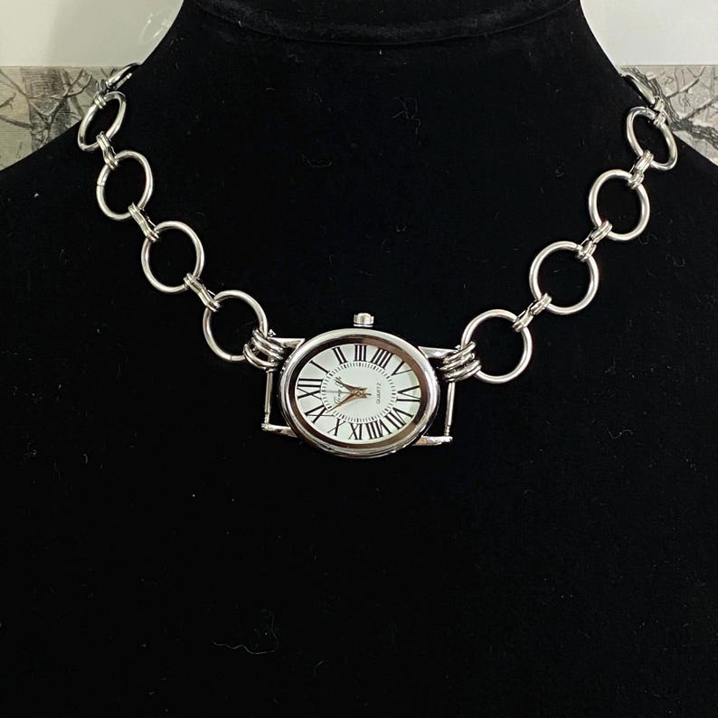 May include: A silver chain necklace with a silver watch attached to it. The watch has a white face with black Roman numeral hour markers and black hands.