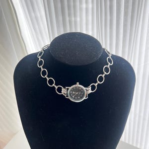 May include: A silver chain necklace with a black watch face attached to it. The watch face has silver hands and black numbers.