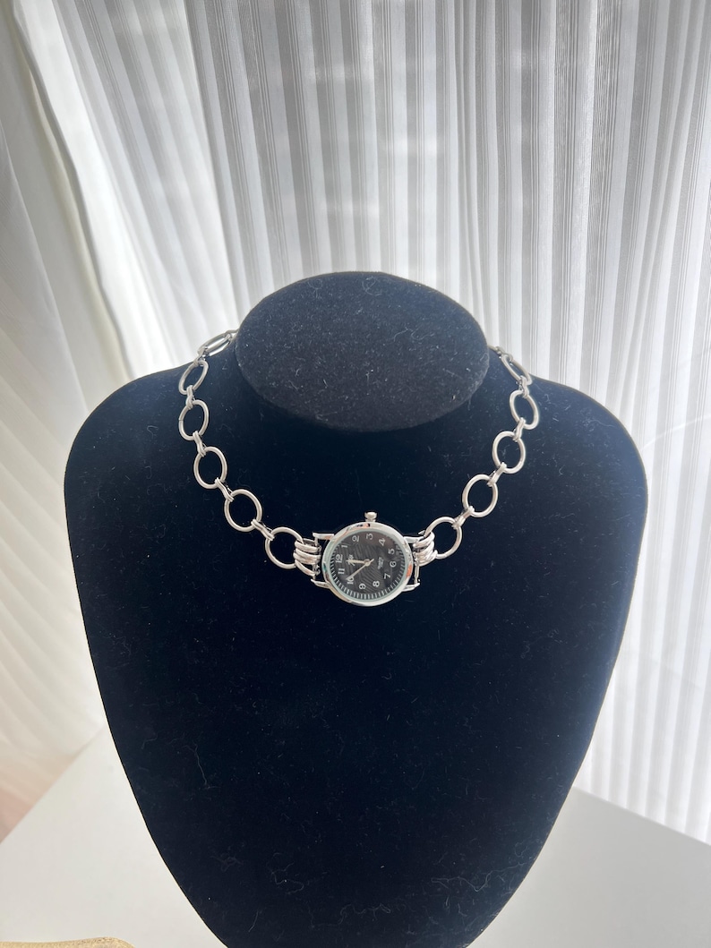 May include: A silver chain necklace with a black watch face attached to it. The watch face has silver hands and black numbers.