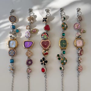 May include: Five decorative watch bracelets with various charms and watch faces. The bracelets feature a mix of floral, heart, and gemstone designs in gold, silver, red, green, and purple. Each bracelet has a chain extension.